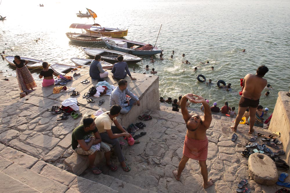 Scène de rue. Street photographie. Un homme s’étire. Des familles jouent dans la rivière Gange. Couleurs. Bénarès. Varanasi. François Guibilato Photographe.